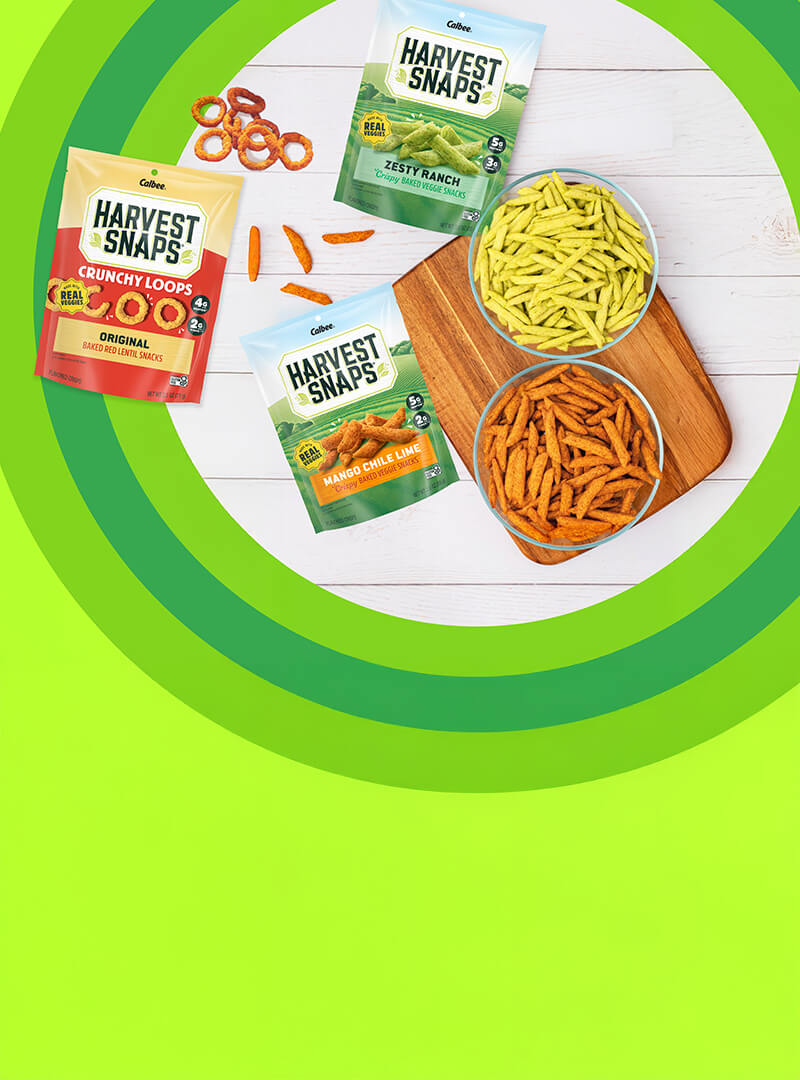 Bags of Harvest Snaps snacks on a wooden surface with bowls of crunchy snacks and a vibrant green background, conveying a fresh, savory theme.